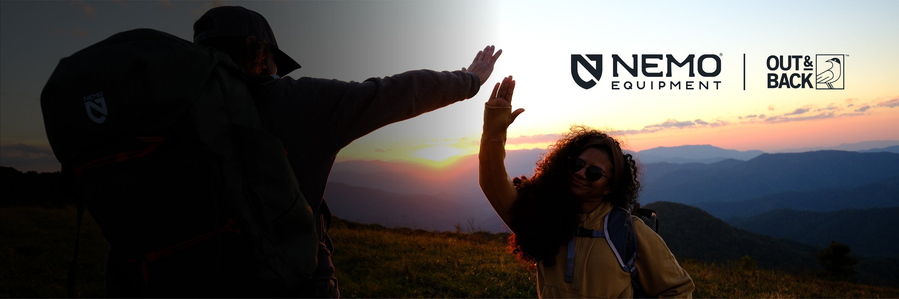 Two people with backpacks high-fiving on a grassy hilltop at sunset, with mountains silhouetted in the background.