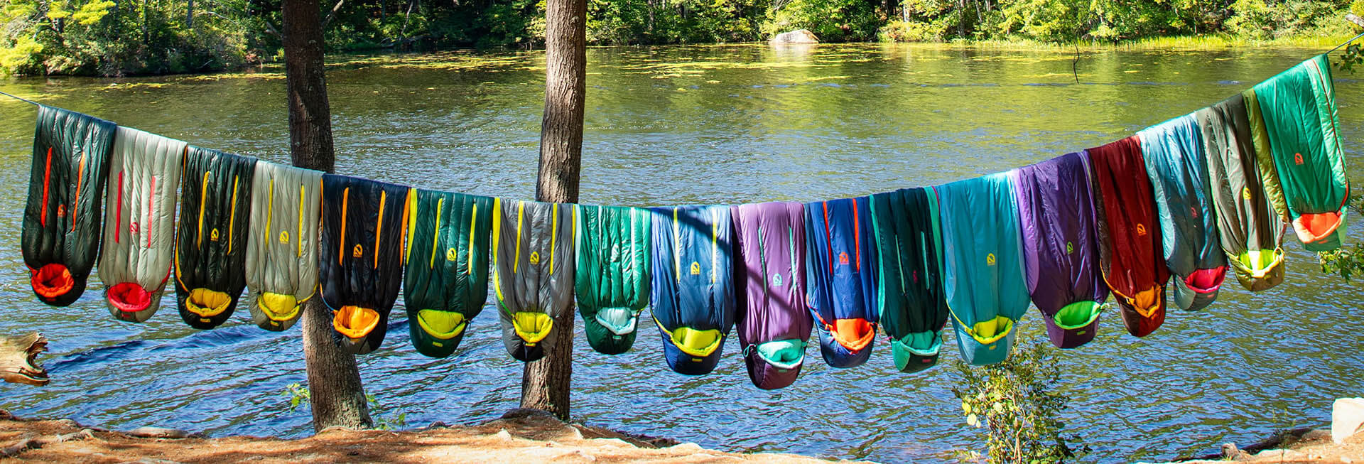 A group of colorful group of sleeping bags hanging on a line near a body of water.